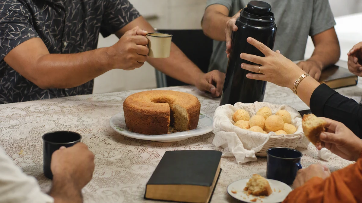Mãos de adultos passando garrafa de café, xícaras, bolo e pão de queijo sobre uma mesa, com uma Bíblia fechada ao lado