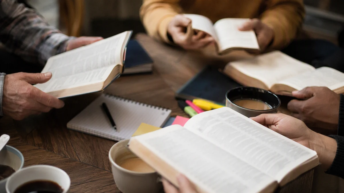 Mãos de adultos segurando Bíblias abertas ao redor de uma mesa de centro, com cadernos, marca-textos e xícaras de café