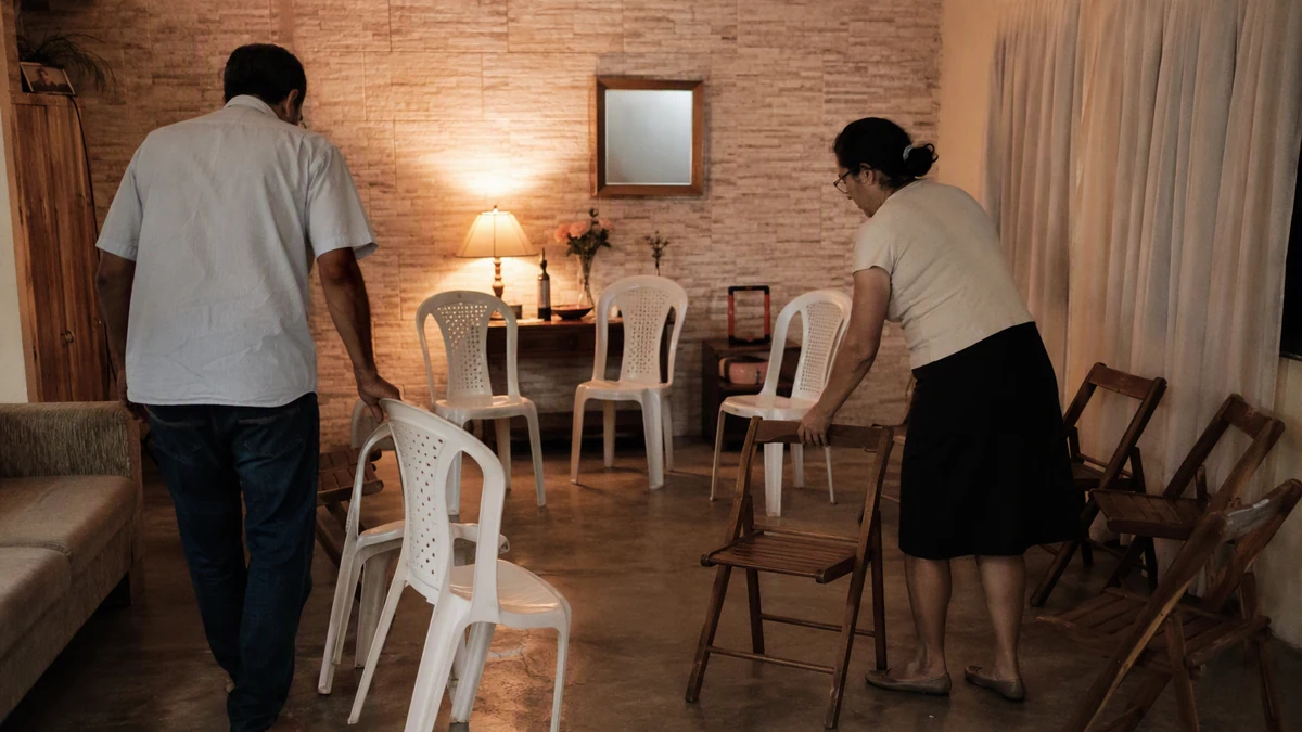 Homem e mulher arrumando cadeiras em círculo numa sala de estar simples, à noite, para uma reunião de pequeno grupo
