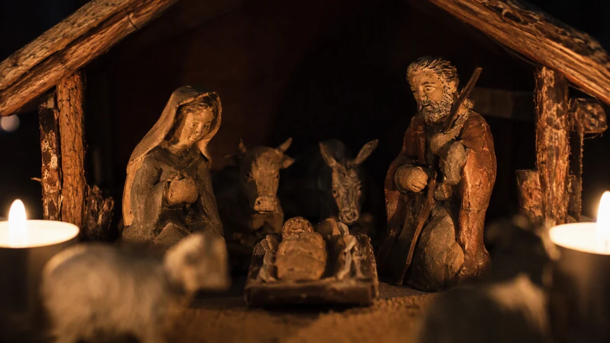 Presépio rústico com figuras de barro dentro de um pequeno estábulo de madeira, iluminado por velas, em fundo escuro