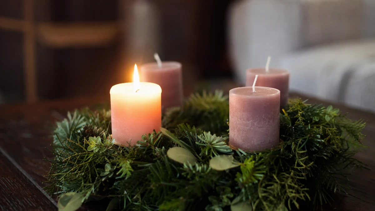Coroa de ramos verdes com velas sobre uma mesa de madeira, uma delas acesa, em luz quente