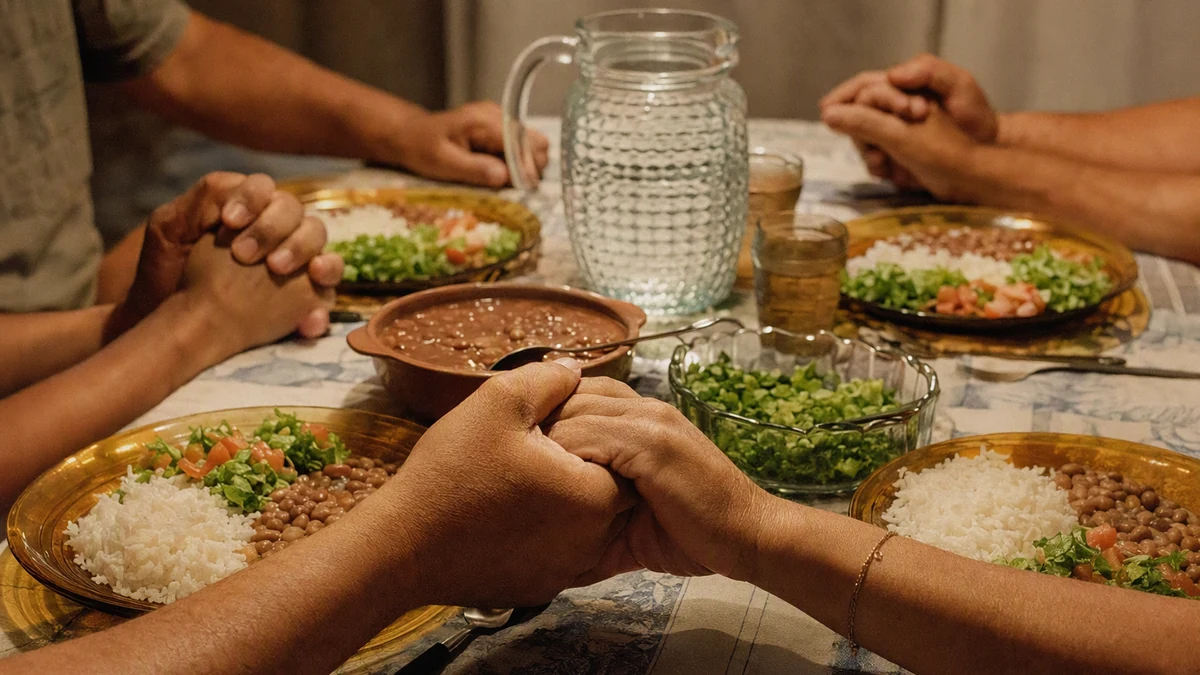 Mãos de adultos unidas em oração ao redor de uma mesa posta com arroz, feijão e salada